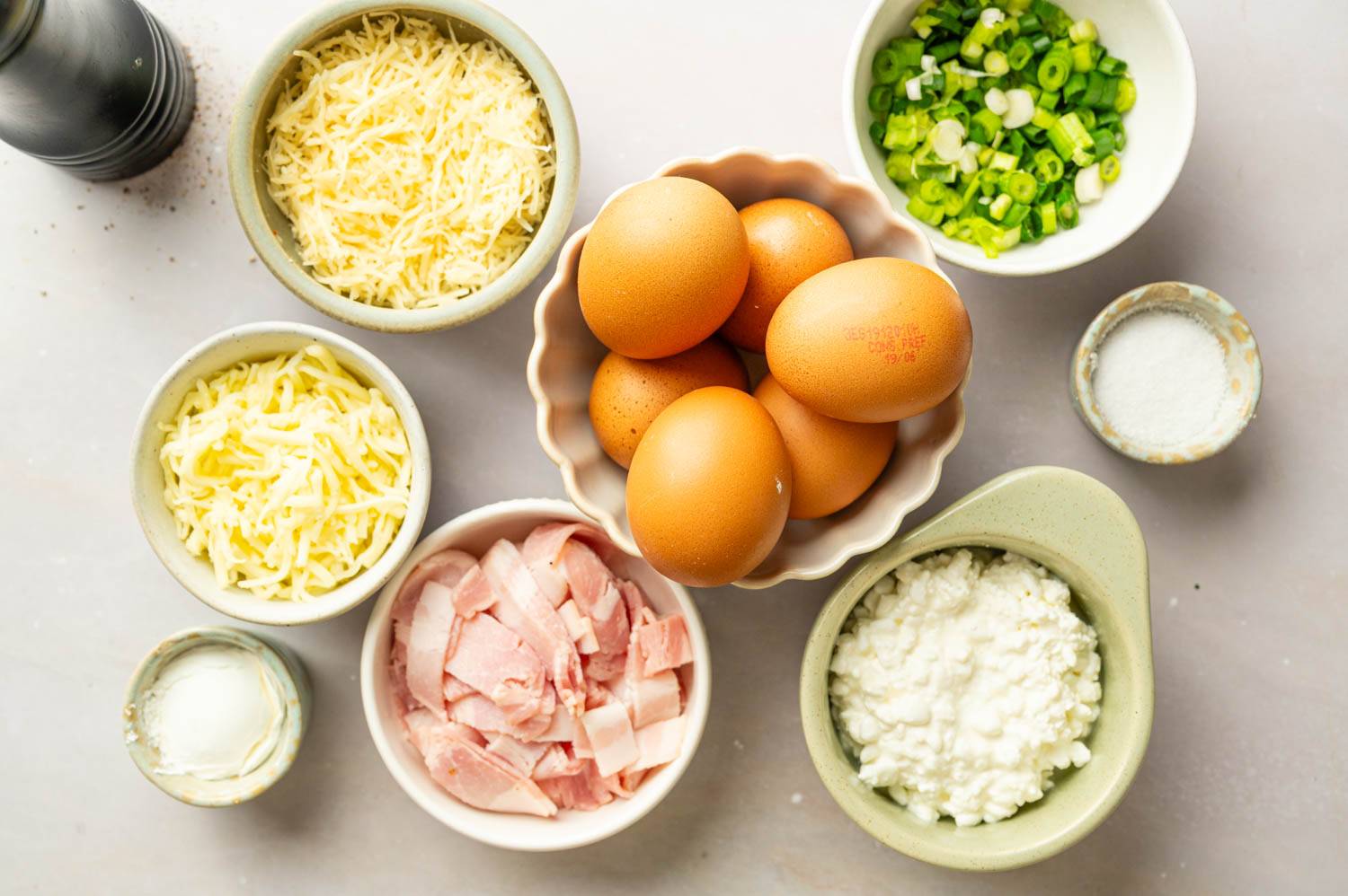 Ingredients for high-protein egg bites arranged on a countertop, including eggs, cottage cheese, Gruyère cheese, mozzarella cheese, chopped turkey bacon, sliced green onions, salt, and pepper.