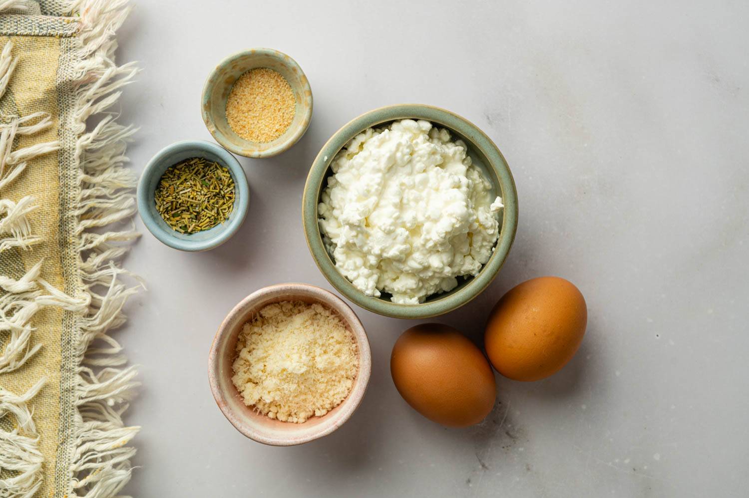 Ingredients for cottage cheese flatbread arranged on a countertop, including cottage cheese, eggs, Parmesan cheese, garlic powder, and Italian seasoning.