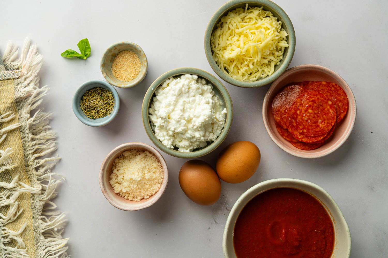 Ingredients for cottage cheese pepperoni pizza arranged in small bowls with eggs, mozzarella cheese, marinara sauce, Parmesan cheese, Italian seasoning, and fresh basil on a gray surface.