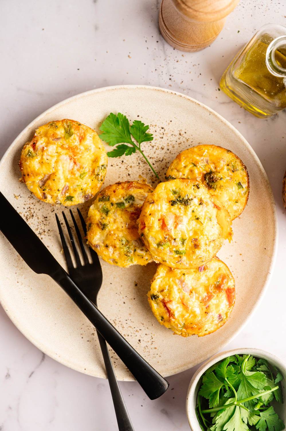 Plate of five golden-brown vegetable muffins with parsley garnish, set on a table with a fork, knife, olive oil, pepper mill, and fresh herbs.