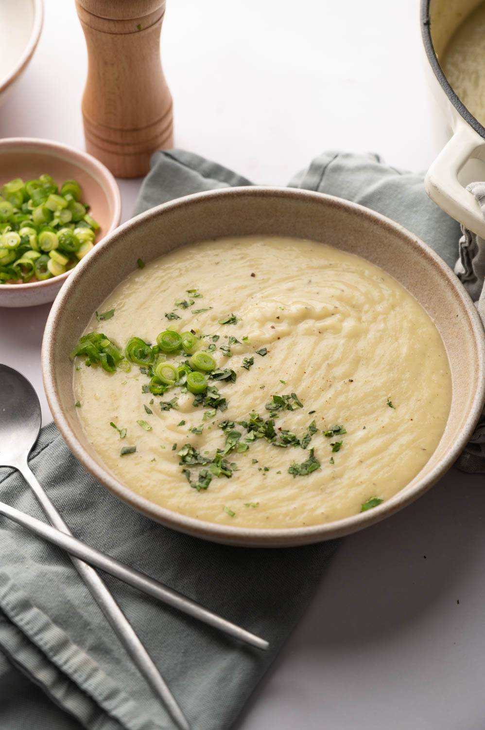 Cream of potato soup in a bowl, garnished with chopped green onions and herbs. Nearby are a pepper mill, green onions and a folded gray napkin with a spoon on it.