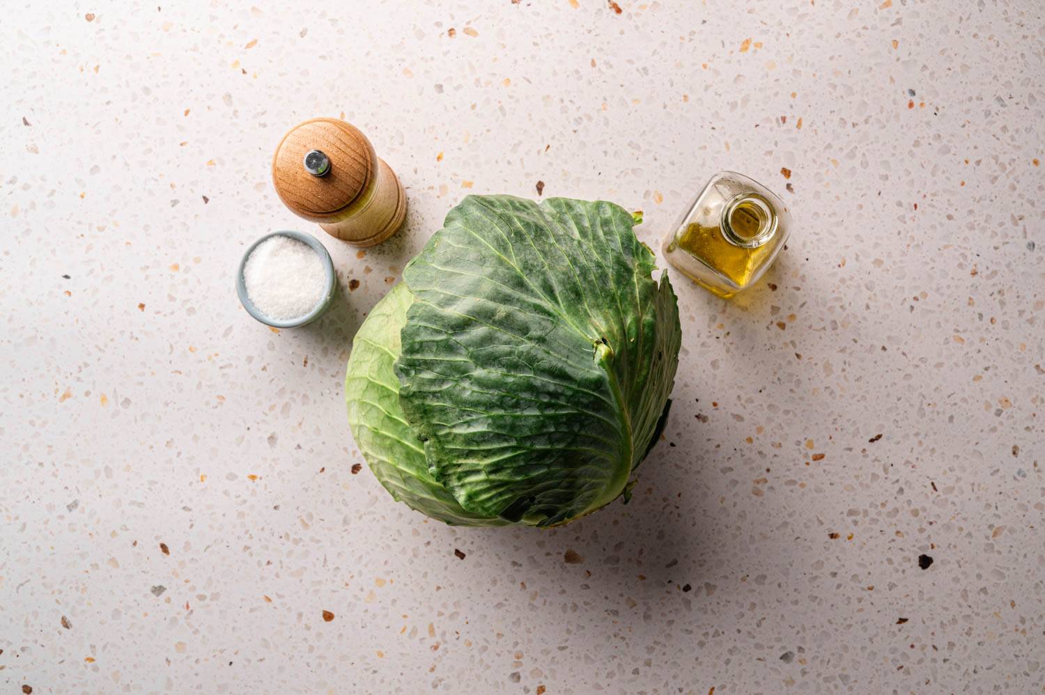 Cabbage on a speckled countertop, surrounded by a wooden pepper grinder, a small bowl of salt, and a glass jar of olive oil.