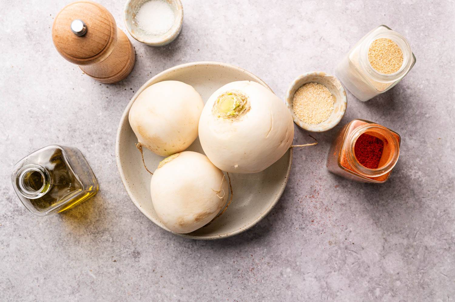 Three radishes on a plate surrounded by spice jars, pepper grinder and olive oil bottle on gray surface.