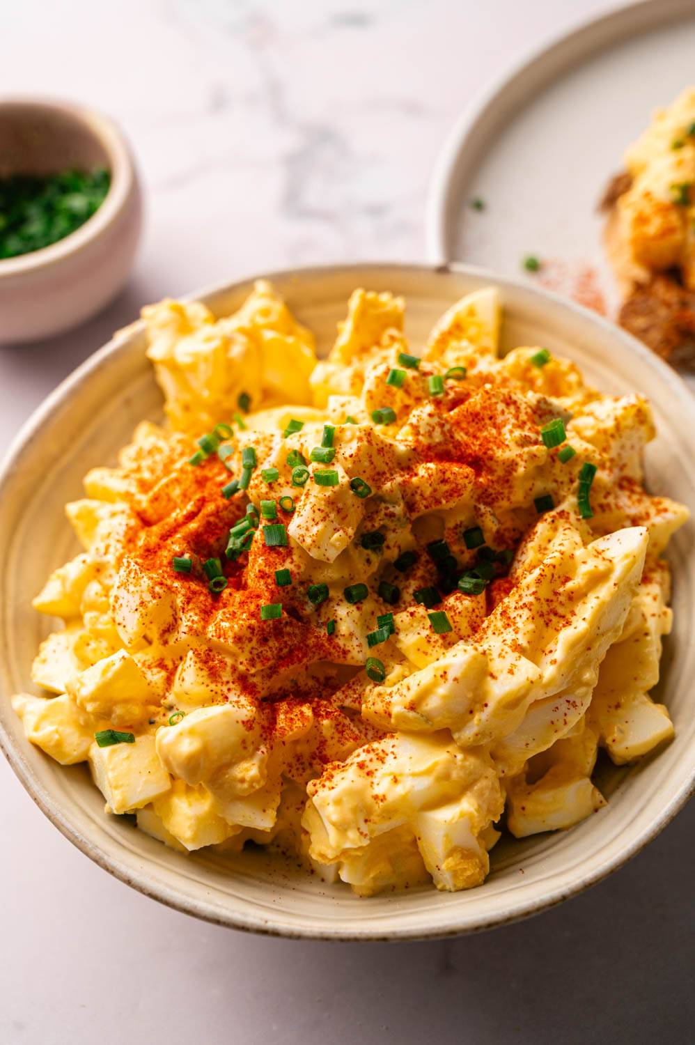 A creamy egg salad is served in a white bowl, topped with chopped chives and paprika for garnish. 