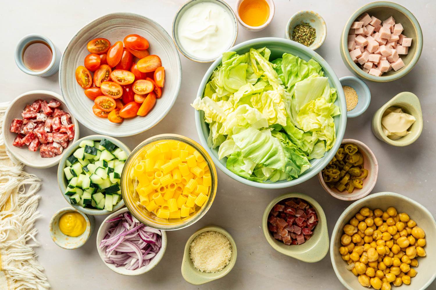 Italian chopped pasta salad ingredients arranged on a countertop, including ditalini pasta, lettuce, chickpeas, tomatoes, cucumbers, deli meats, Parmesan cheese, and dressing ingredients.