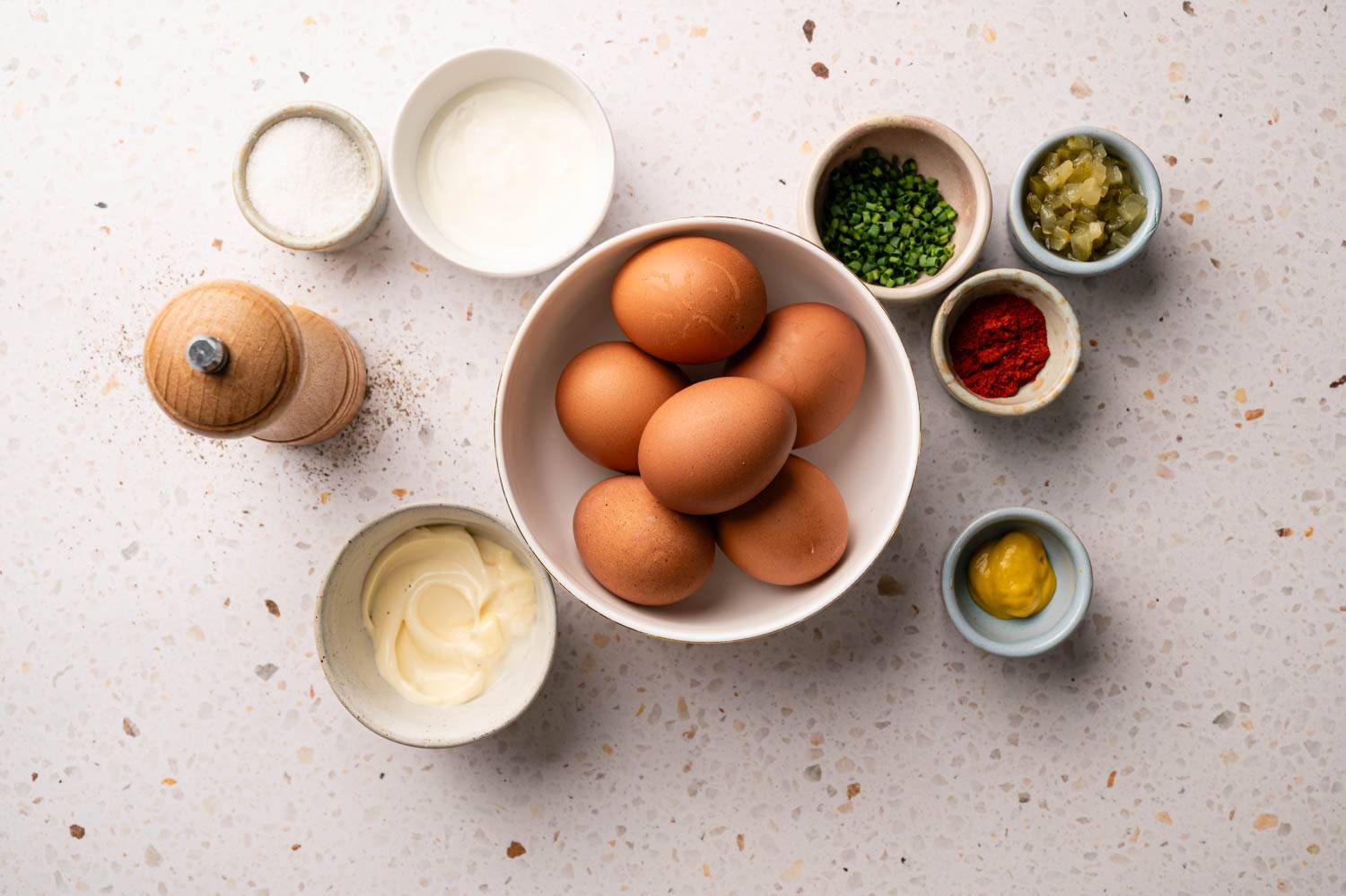 A bowl of brown eggs sat on the white countertop, surrounded by bowls of salt, yogurt, chives, seasonings, paprika, mustard and mayonnaise, and a pepper grinder.