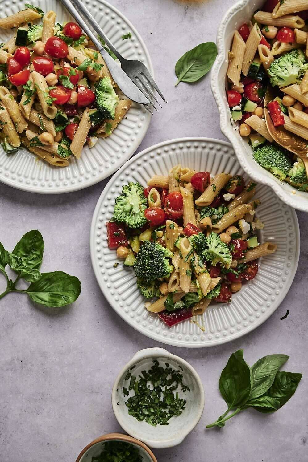 A bowl of vibrant pasta salad with cherry tomatoes, broccoli and chickpeas, decorated with fresh herbs on the texture.