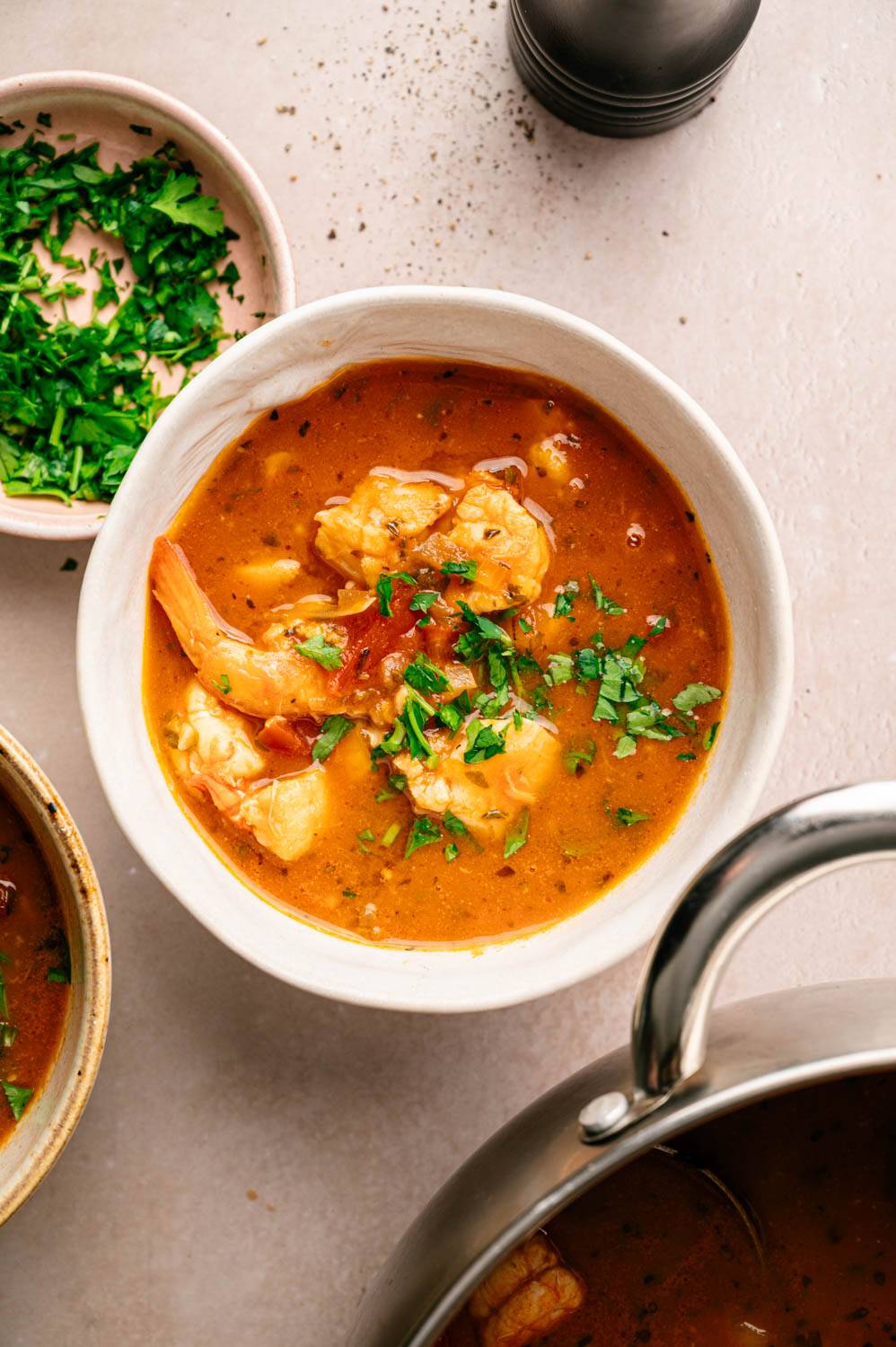 A bowl of shrimp bisque with fresh parsley. A rich orange broth with shrimp chips surrounded by a small plate of parsley and pepper grinders.