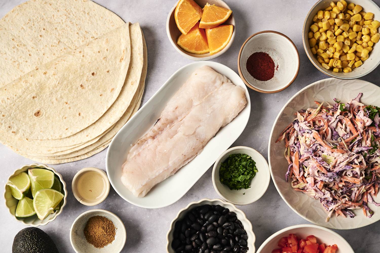 The ingredients of the tacos are arranged on the countertops: tortillas, fillets, coleslaw, lime, avocado, corn, black beans and citrus.