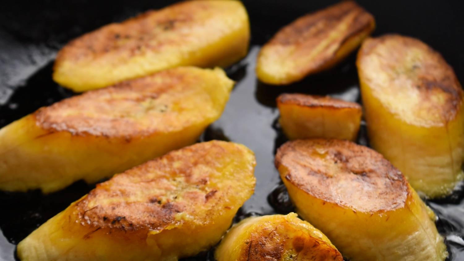 Sliced ripe plantains frying in a pan, golden brown with crispy edges, creating a warm and appetizing scene. The surface glistens with oil.