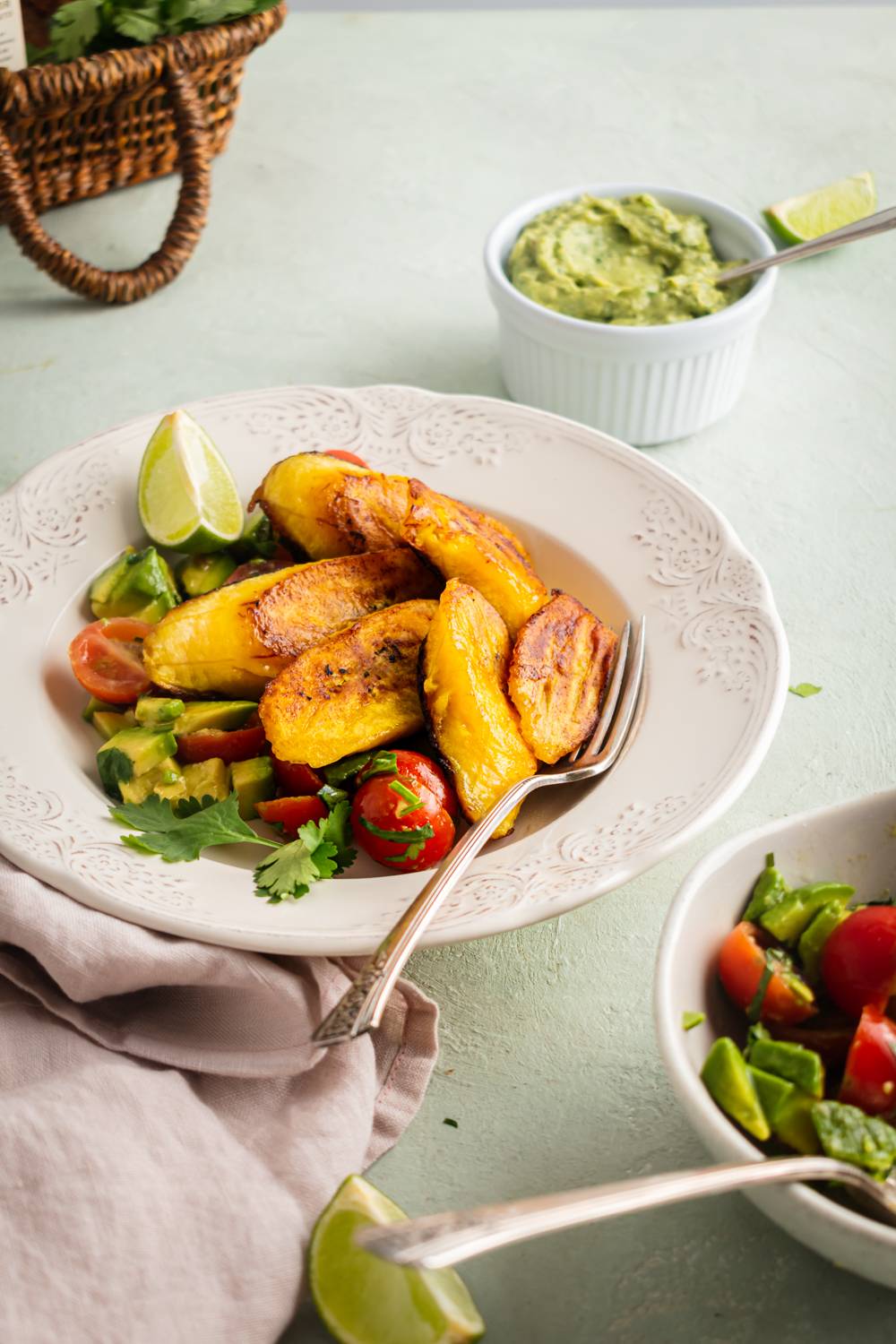 A plate of golden fried plantains garnished with lime sits atop a salad of cherry tomatoes and avocado, accompanied by a bowl of guacamole.