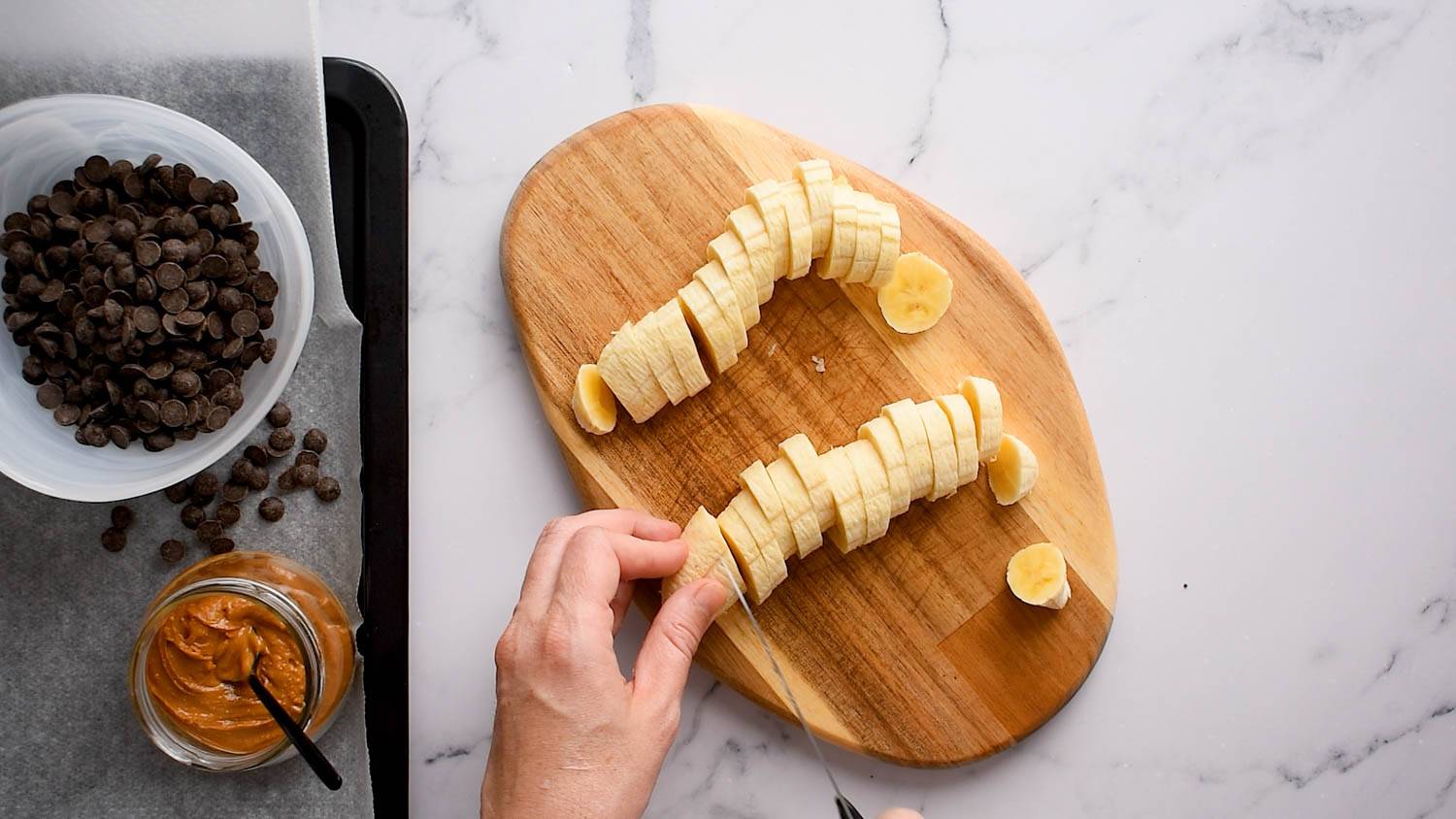 Cut a pile of banana pieces on a wooden cutting board with one hand around chocolate chips and peanut butter.
