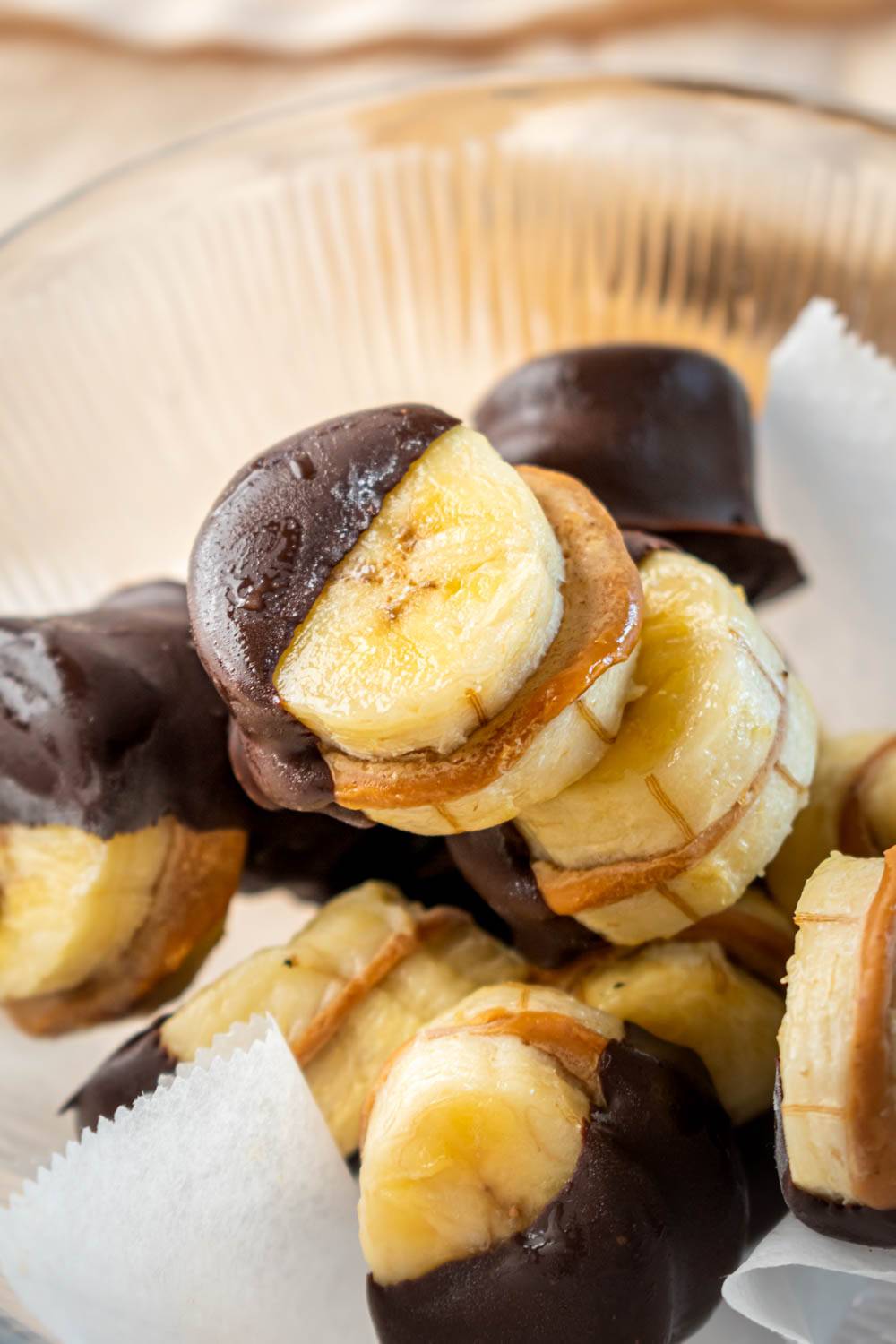 Close-up knife of thinly sliced ​​banana dipped in chocolate and placed in a glass bowl with white paper lining.