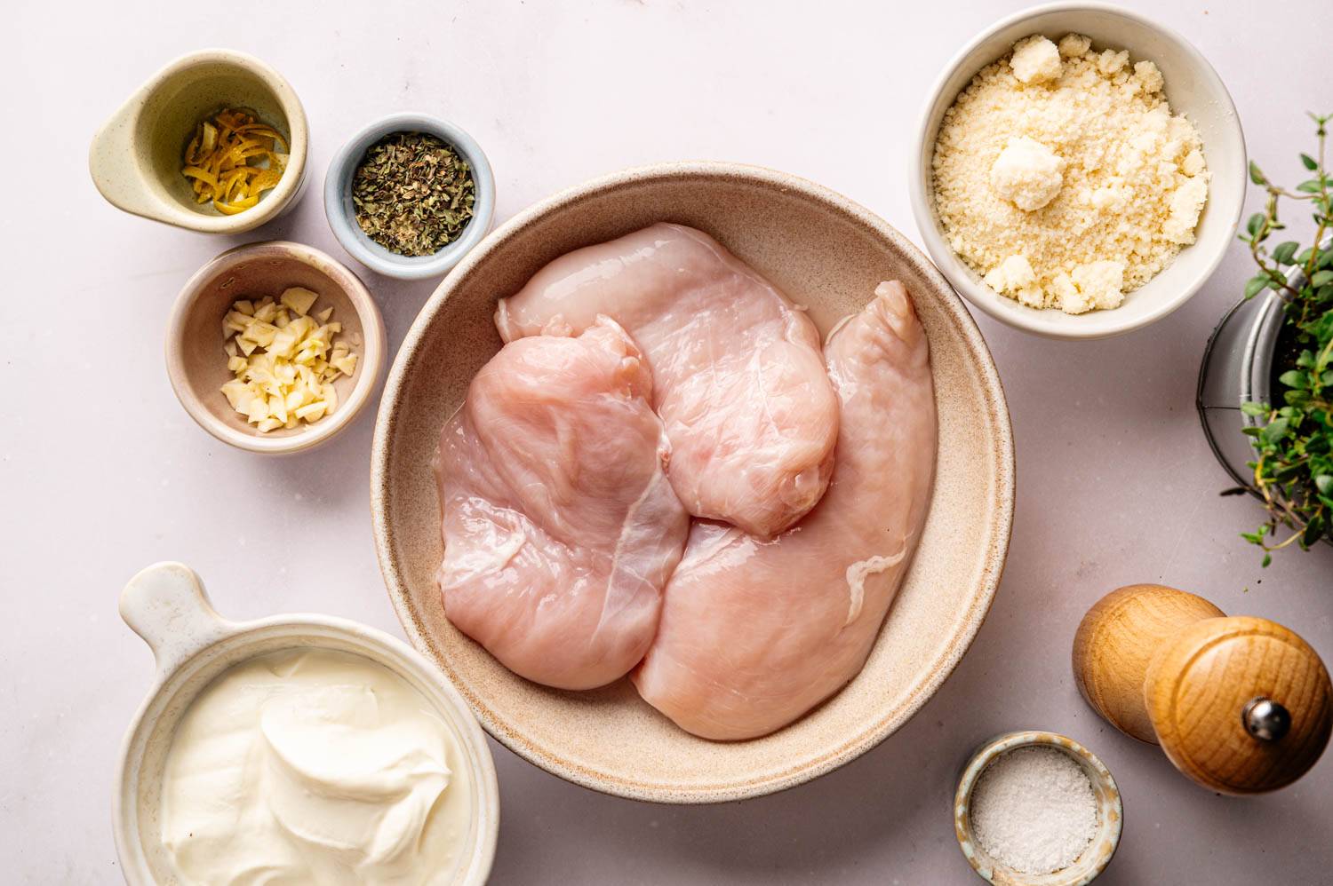 Fresh raw chicken breast in a bowl with garlic, dried herbs, lemon zest, Greek yogurt, grated Parmesan cheese, salt and pepper.
