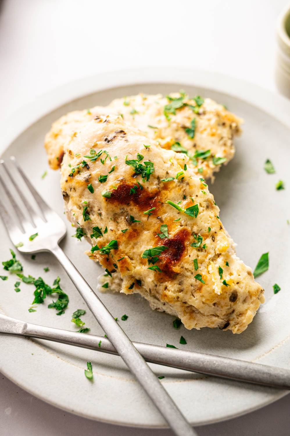Close-up of golden cheesy grilled chicken breast with fresh parsley on plate.