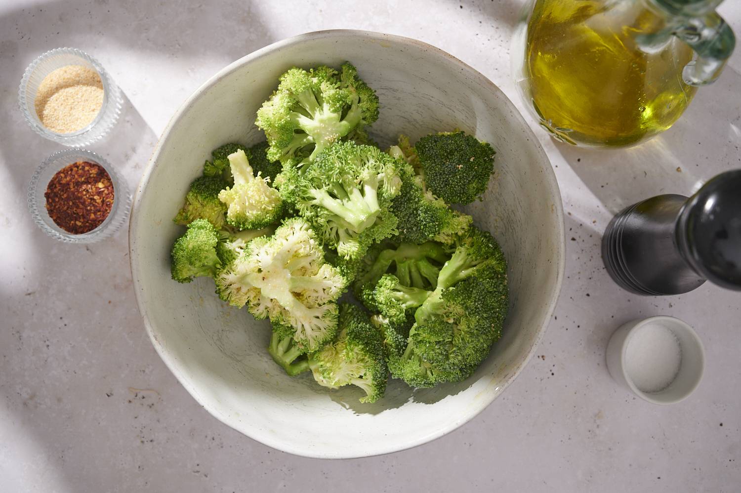 A bowl of fresh broccoli, surrounded by olive oil, salt and spices on a light countertop.