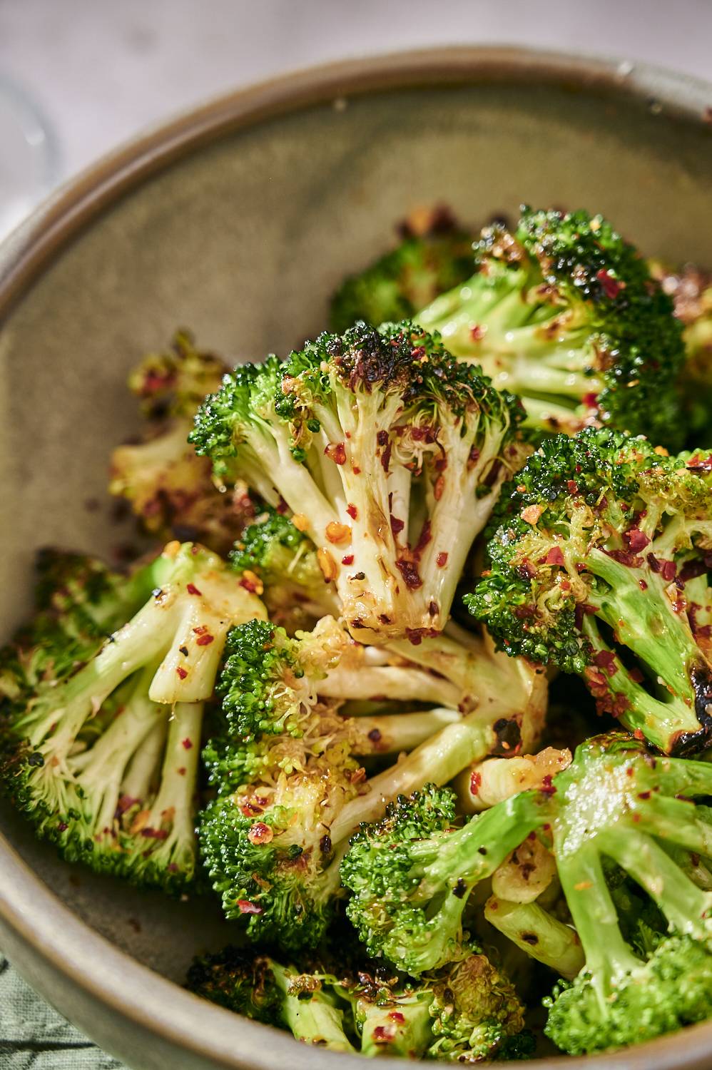 Close-up of small roasted broccoli flowers seasoned with chili flakes in textured bowl on light background.