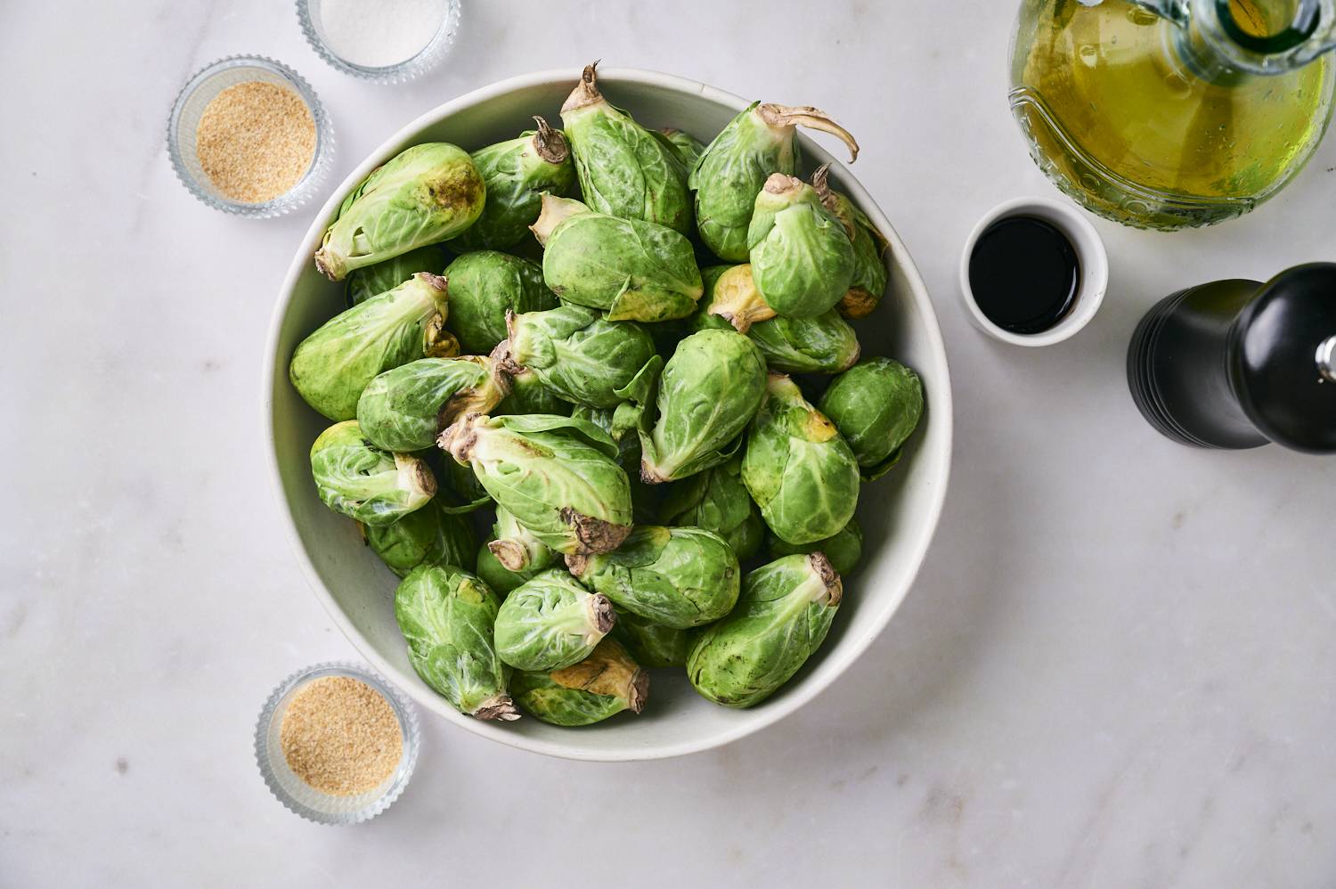 Fresh Brussels fresh Brussels sprouts in a white bowl surrounded by olive oil, balsamic vinegar, garlic powder, salt and pepper grinders that can be prepared for grilling.