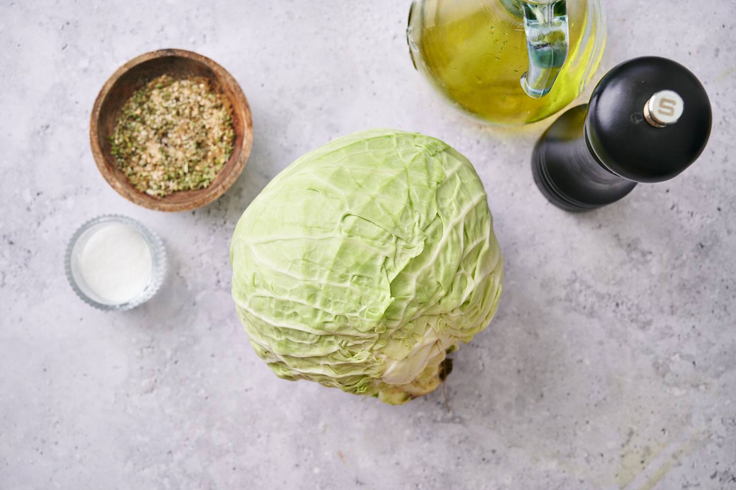 Fresh green cabbage sits on a light-colored surface next to a bowl of seasoning, salt, olive oil and pepper grinder.