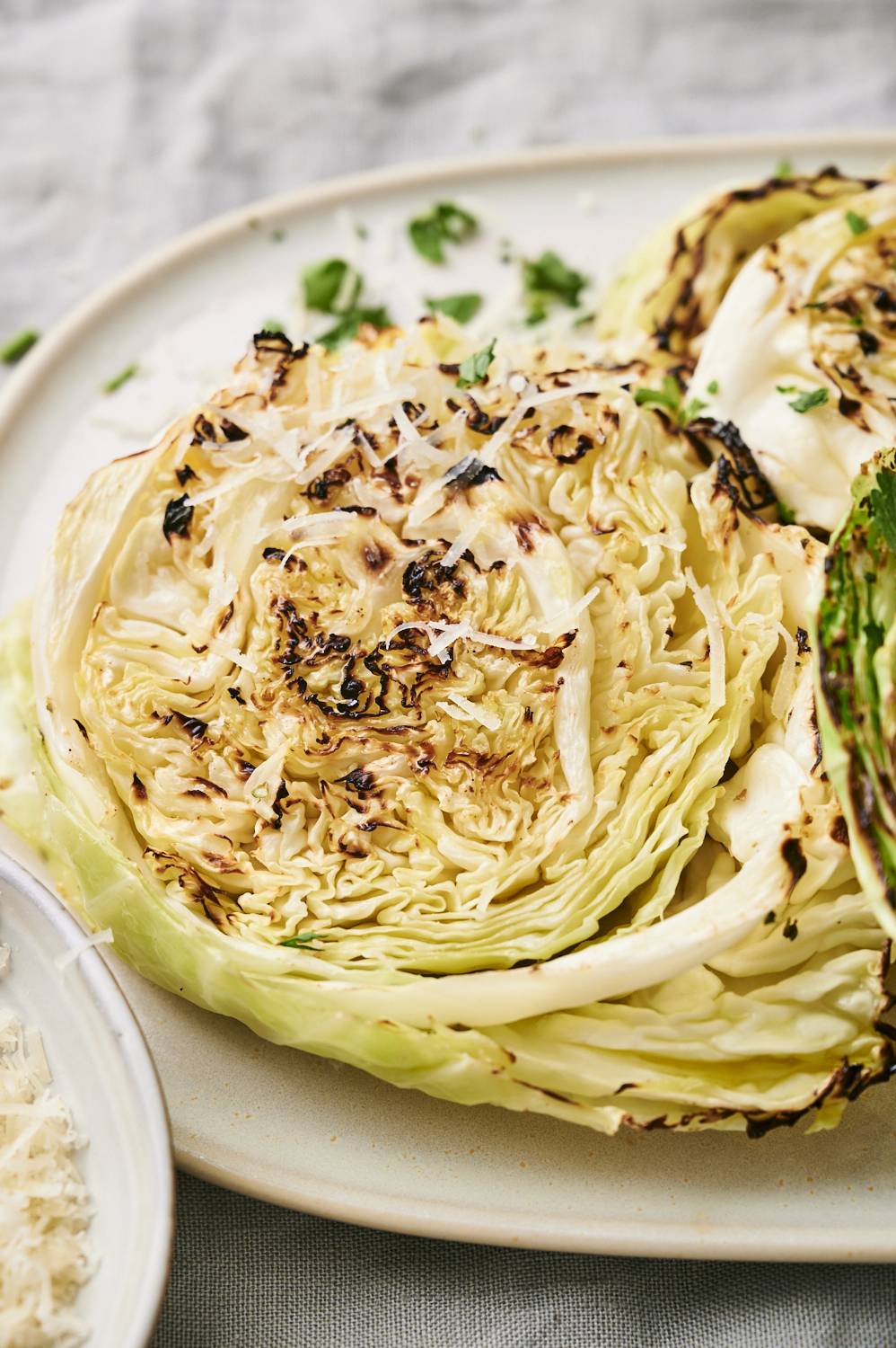 Roasted cabbage wedges on beige plate with grated cheese and green herbs, sit on a light grey background in texture.