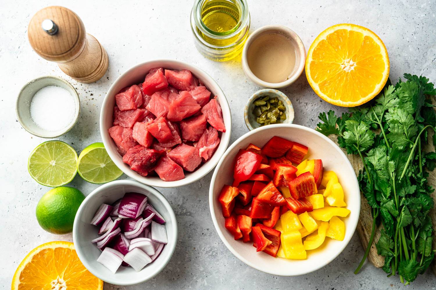 Overhead shot of raw ingredients for carne asada skewers arranged on a light gray surface, including a bowl of cubed steak, a bowl of red and yellow bell pepper chunks, a bowl of diced red onion, halved limes, a halved orange, a small dish of diced jalapeno, fresh cilantro sprigs, a jar of olive oil, a small bowl of vinegar, plus salt and a pepper grinder.