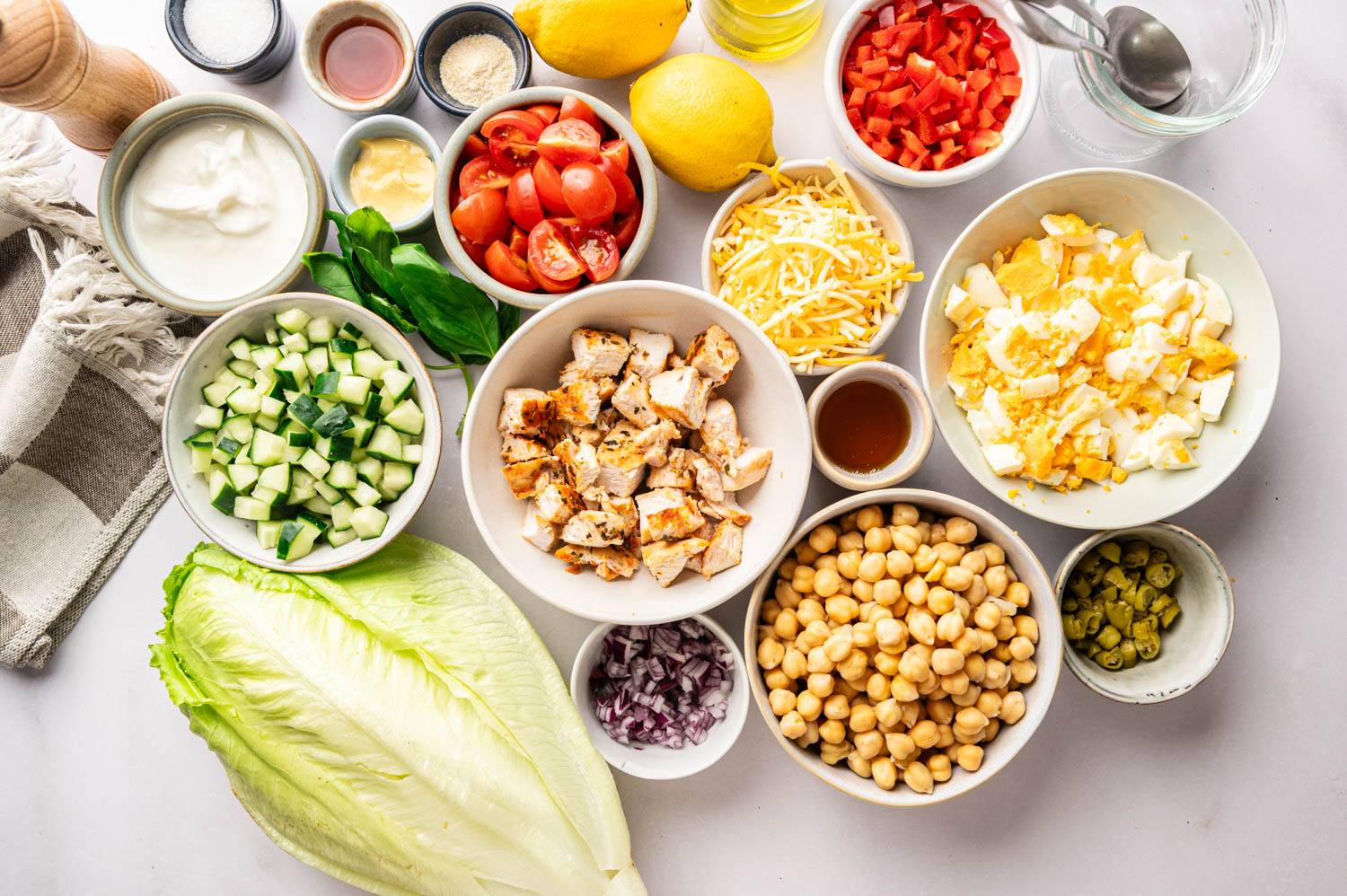 Overhead shot of chopped salad ingredients in prep bowls, including diced cucumber, cherry tomatoes, red bell pepper, chopped hard-boiled eggs, shredded cheese, chickpeas, diced grilled chicken, red onion, sliced pepperoncini, romaine lettuce, lemons, and small bowls of dressing ingredients on a marble countertop.