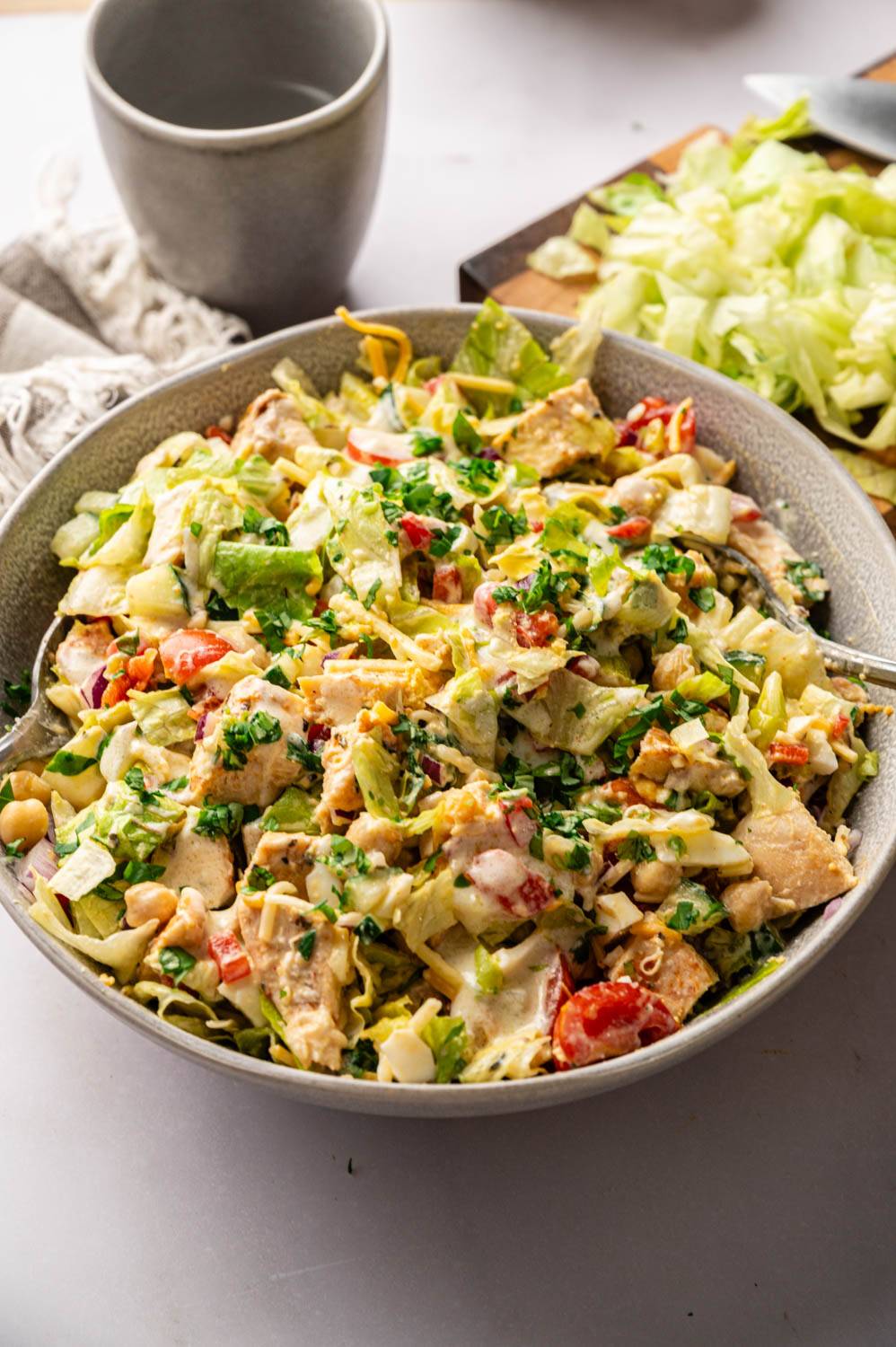Close-up of grilled chicken chopped salad in a stoneware bowl with two forks, tossed with creamy dressing and topped with fresh parsley, next to a gray mug and a cutting board of shredded lettuce.