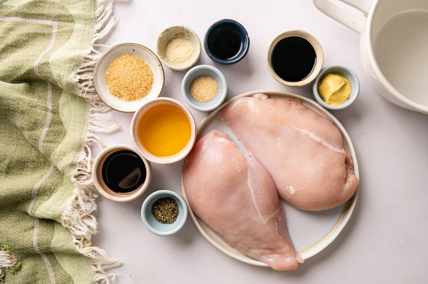 Two raw chicken breasts on a plate surrounded by small bowls of soy sauce, balsamic vinegar, honey, Dijon mustard, garlic powder, brown sugar, and Italian seasoning.