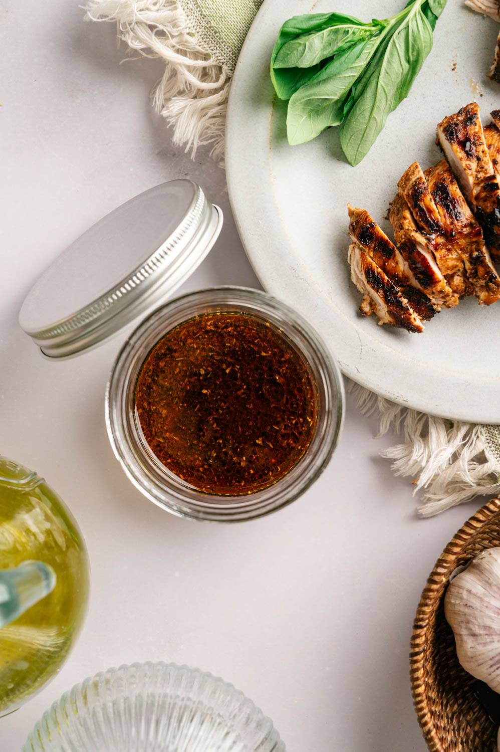 A glass jar of chicken marinade with visible herbs and spices, set beside a plate of grilled and sliced chicken garnished with fresh basil.