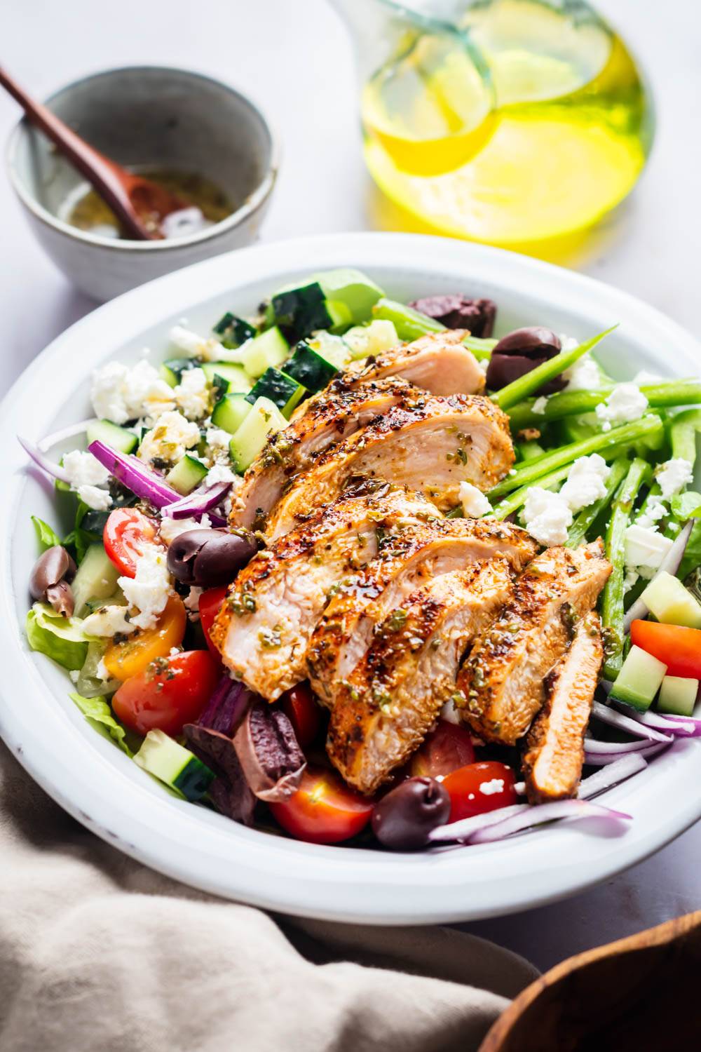 A vibrant salad with grilled chicken slices, tomatoes, cucumbers, olives, red onions, and feta cheese on a white plate. Olive oil and dressing are nearby.