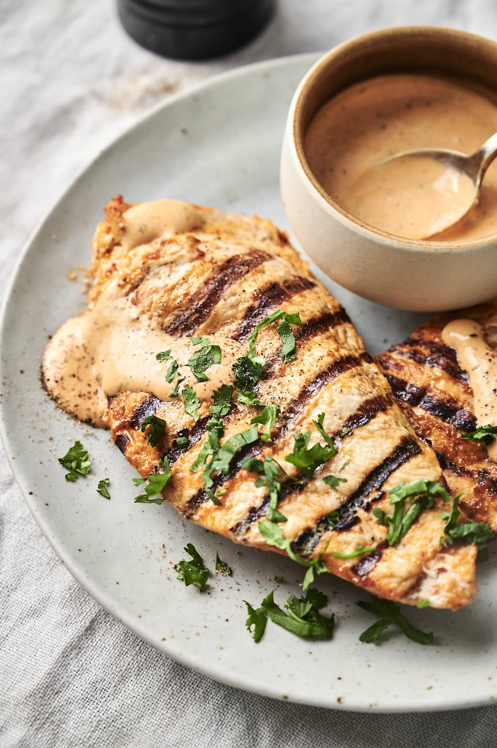 Grilled chicken breasts with char marks, garnished with fresh parsley on a plate. A creamy sauce is served in a bowl beside them.