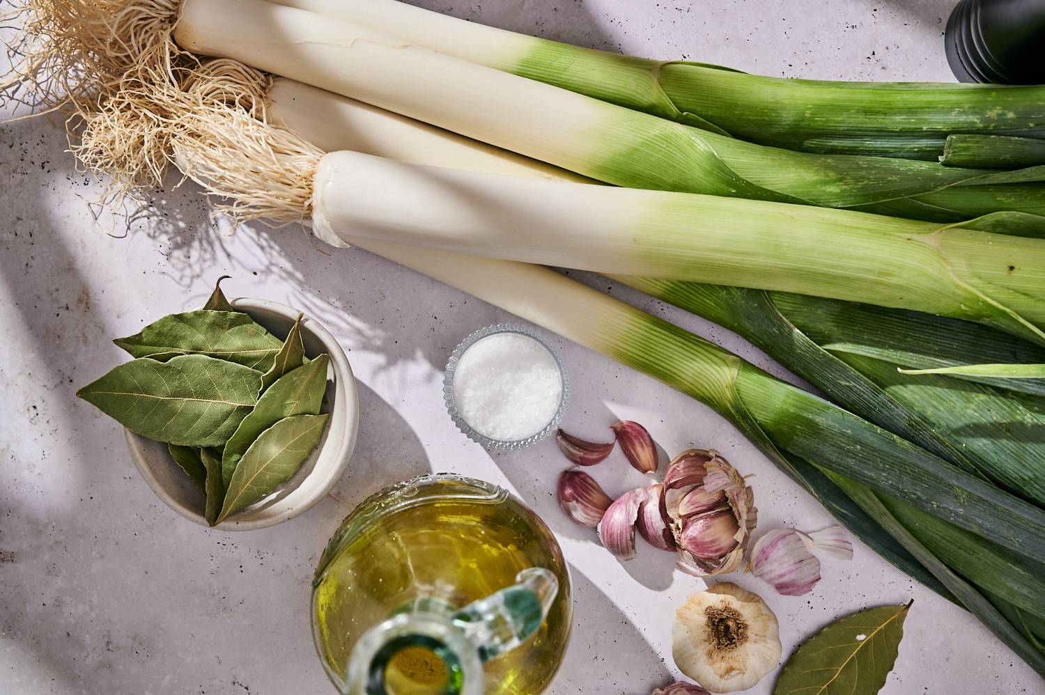 Fresh leeks, fresh leeks with garlic, bay leaves, olive oil and salt, arranged on a light countertop for grilling.