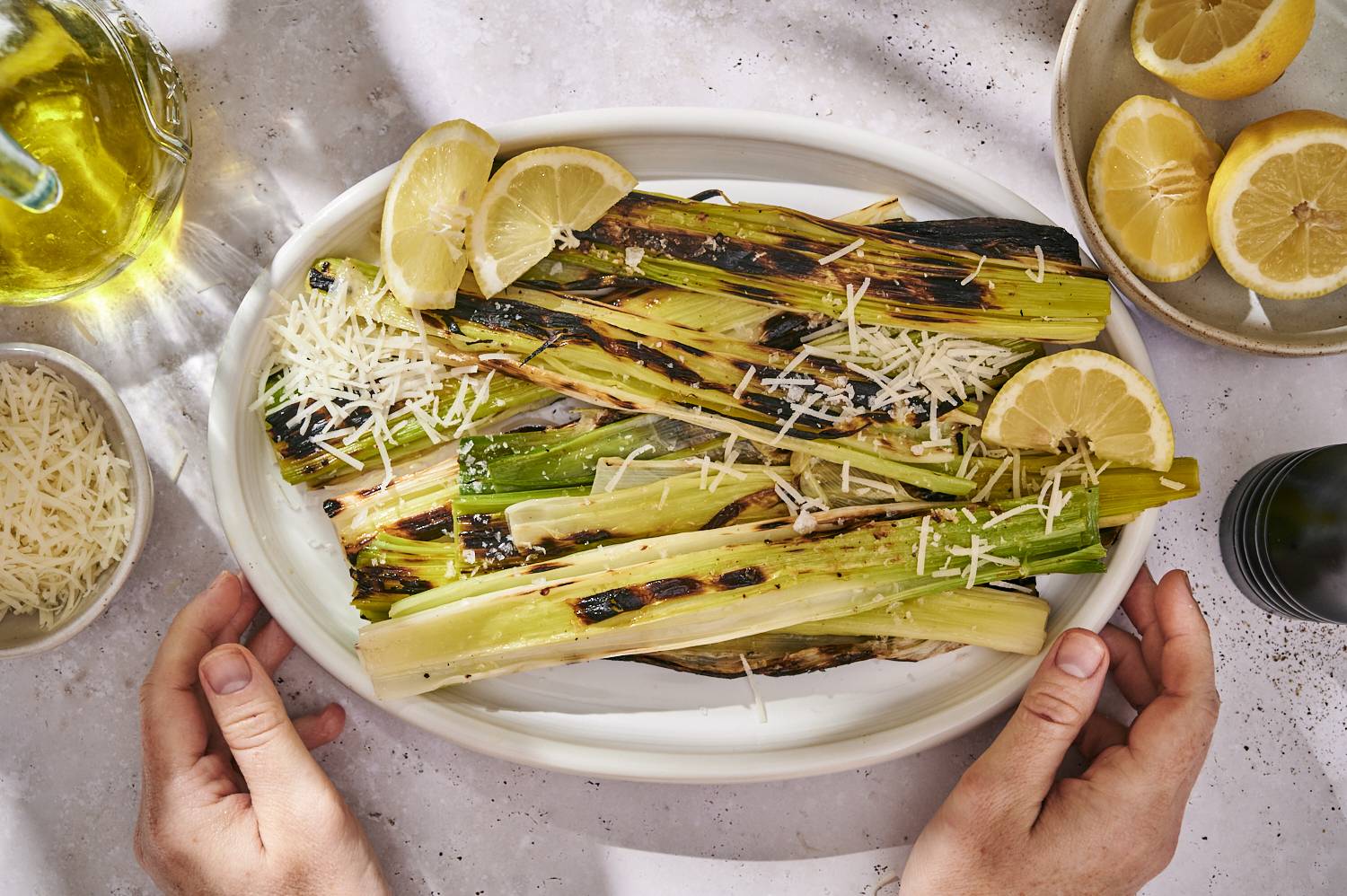 The roasted leeks have charcoal marks on top with grated parmesan cheese and served with lemon wedges on a white plate with some olive oil nearby.