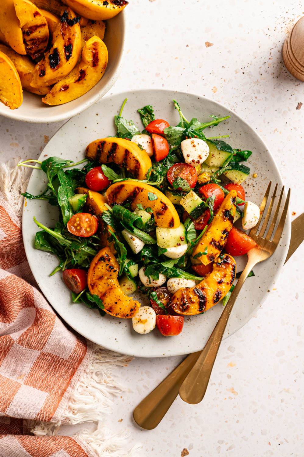 Grilled peach salad on a light gray plate with arugula, halved cherry tomatoes, mozzarella pearls, and charred peach slices, served with gold flatware and an orange checkered napkin.