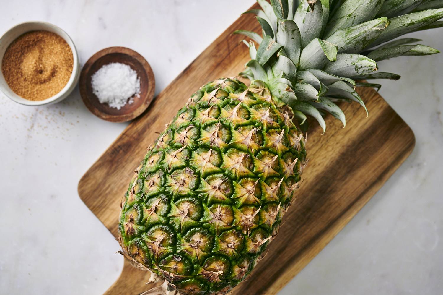 Fresh pineapple on wooden cutting board accompanied by a bowl of brown sugar and sea salt.
