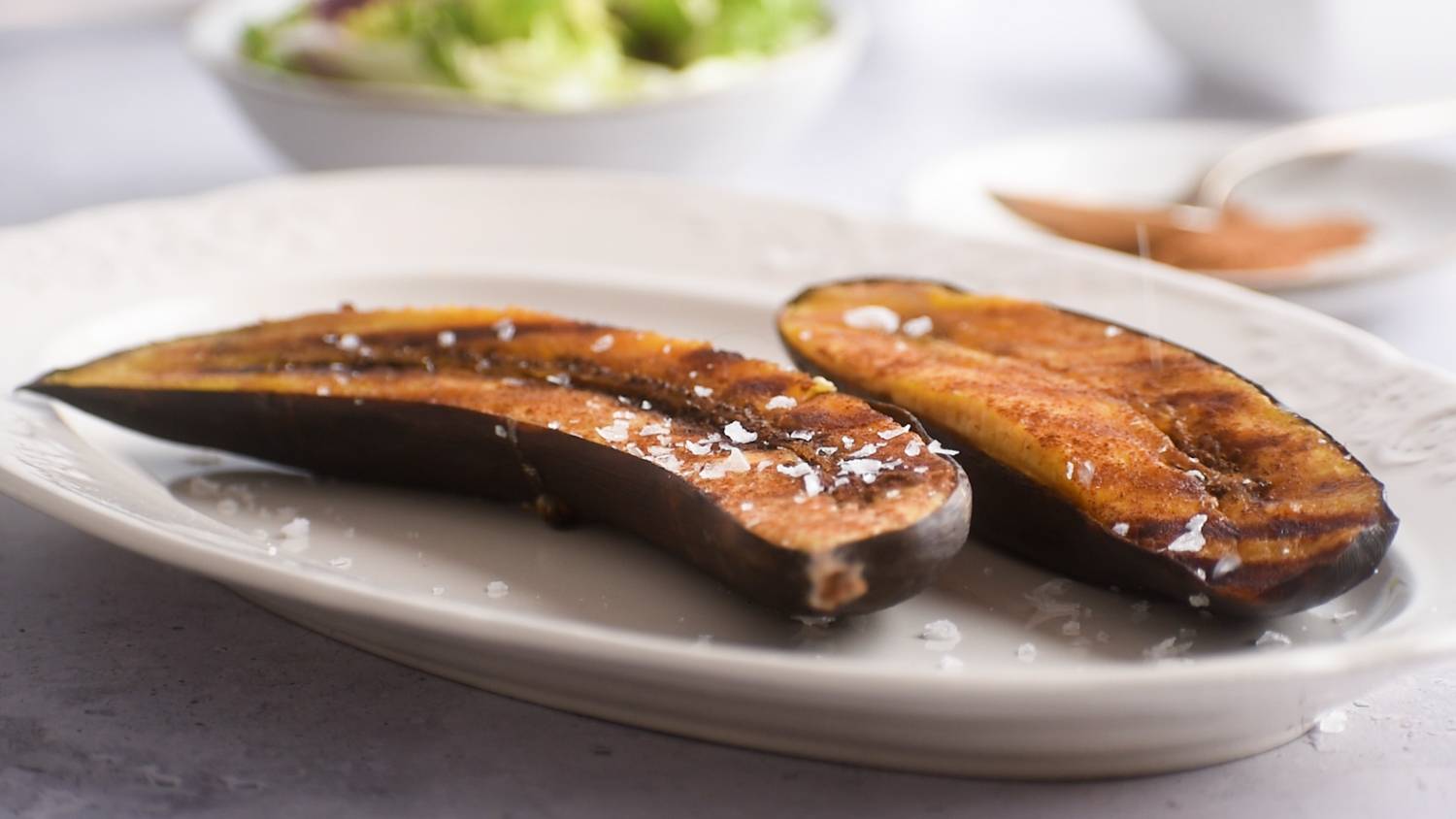 Two baked plantains seasoned with salt, placed on a decorative plate with fresh salad on the background.