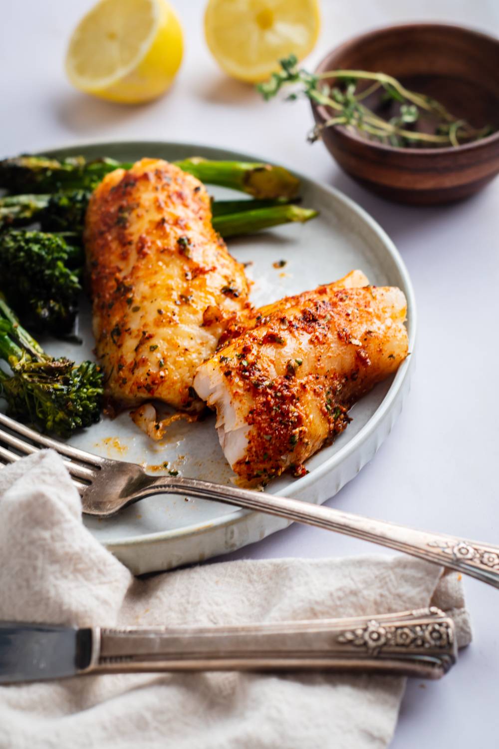 A plate of seasoned fillets with toasted broccoli, topped with thyme and accompanied by lemon half and old-fashioned silverware.