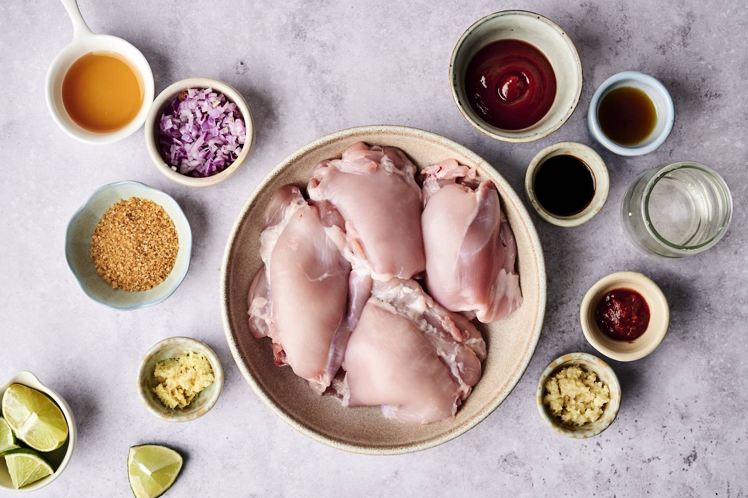 A bowl of raw chicken thighs is surrounded by a variety of ingredients, including chopped onions, seasonings, spices and lemon wedges on the countertop.