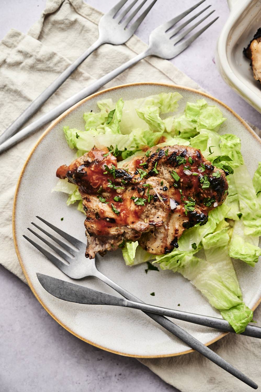 Grilled chicken thighs sprinkled with sauce and herbs topped on the lettuce bed, accompanied by silver cutlery on the textured table.