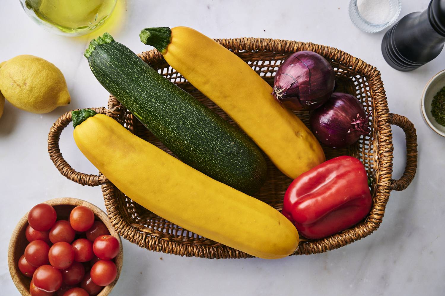 A basket filled with fresh zucchini, yellow pumpkin, red bell peppers and red onions surrounded by cherry tomatoes and lemons.