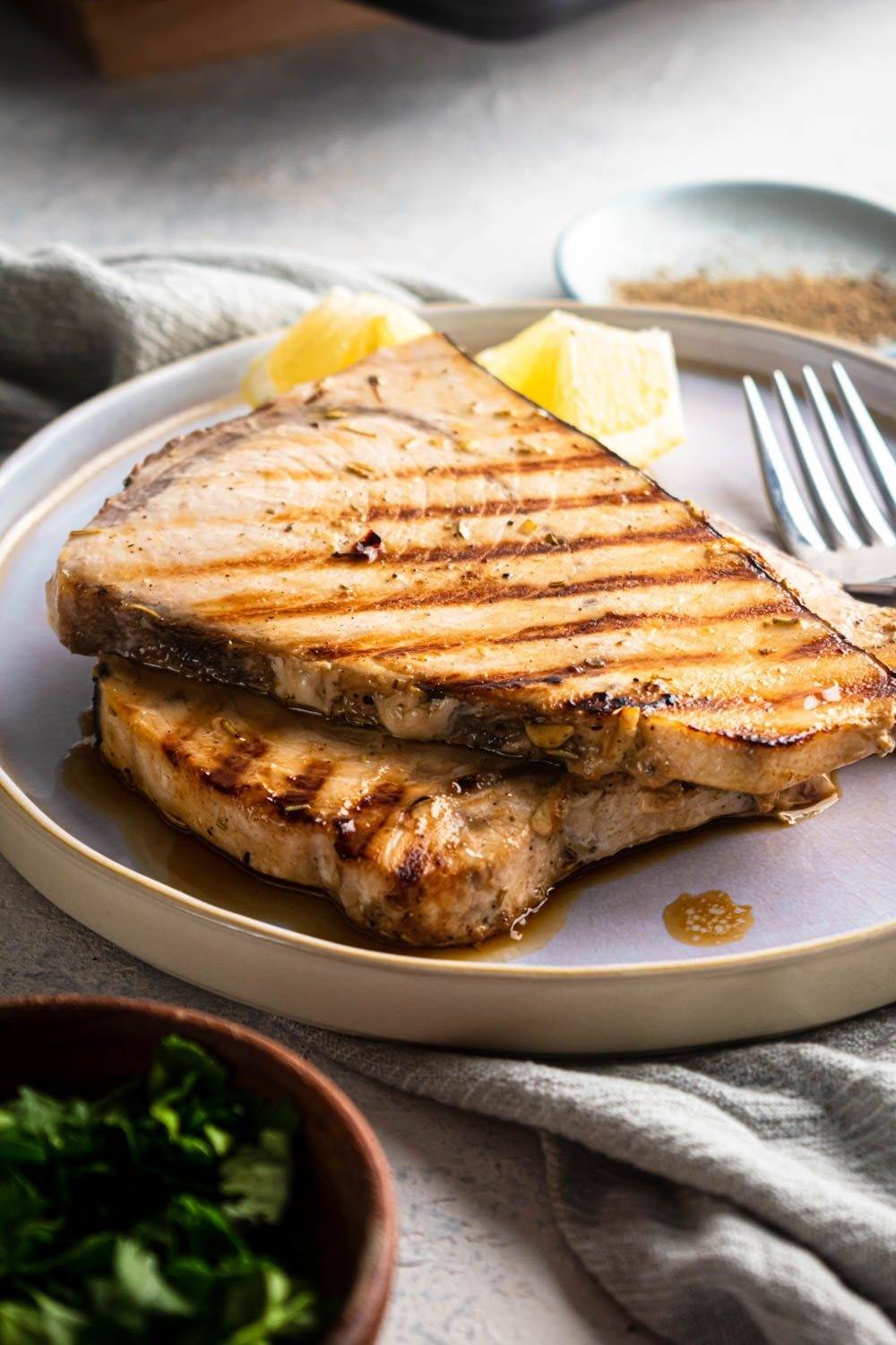 Swordfish are roasted on a plate with herbs and garlic, with fresh lemon on the side.