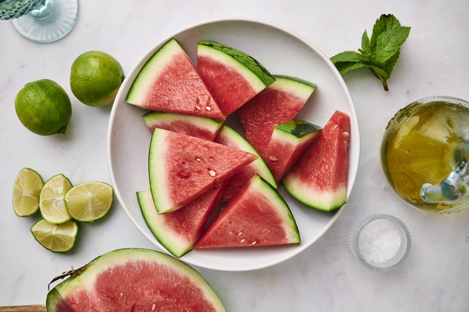 Ingredients for grilled watermelon including fresh sliced watermelon, limes, olive oil, salt, and mint.