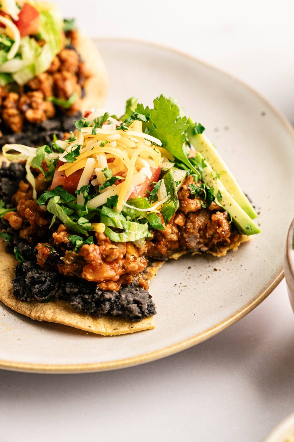 Ground turkey tostada topped with black beans, seasoned turkey, shredded lettuce, diced tomato, shredded cheese, avocado slices, and fresh cilantro on a ceramic plate.