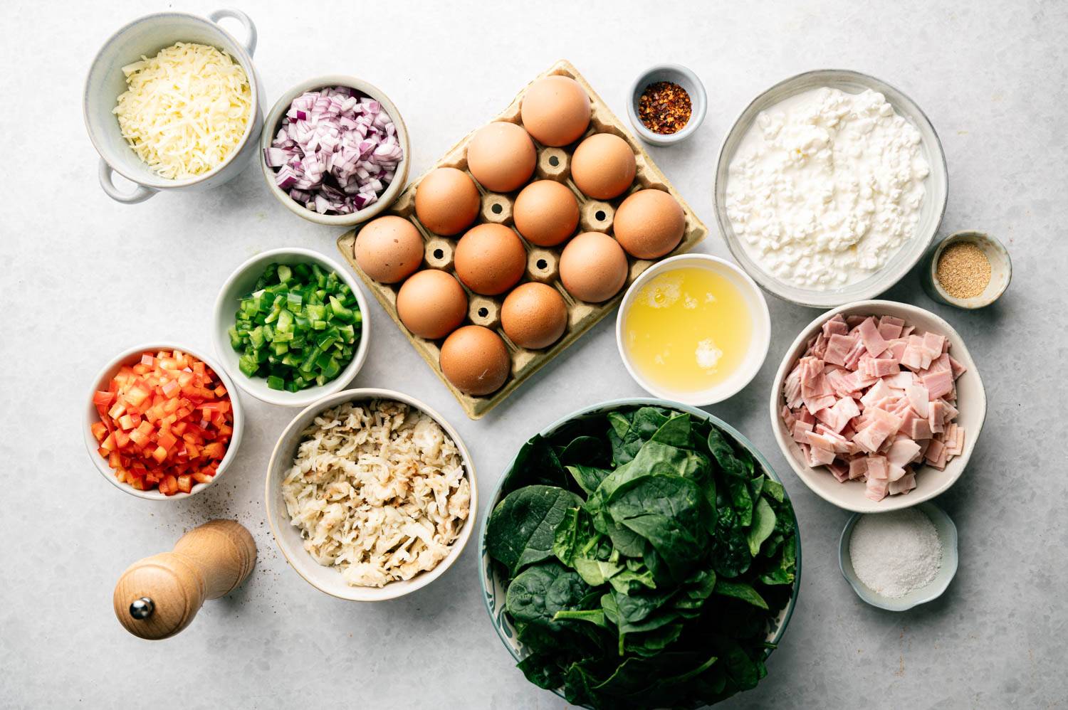 Selected ingredients including eggs, spinach, diced ham, cheese, chopped onions, green peppers, mozzarella and bread crumbs on a white surface evoke a sense of freshness and readiness to cook.