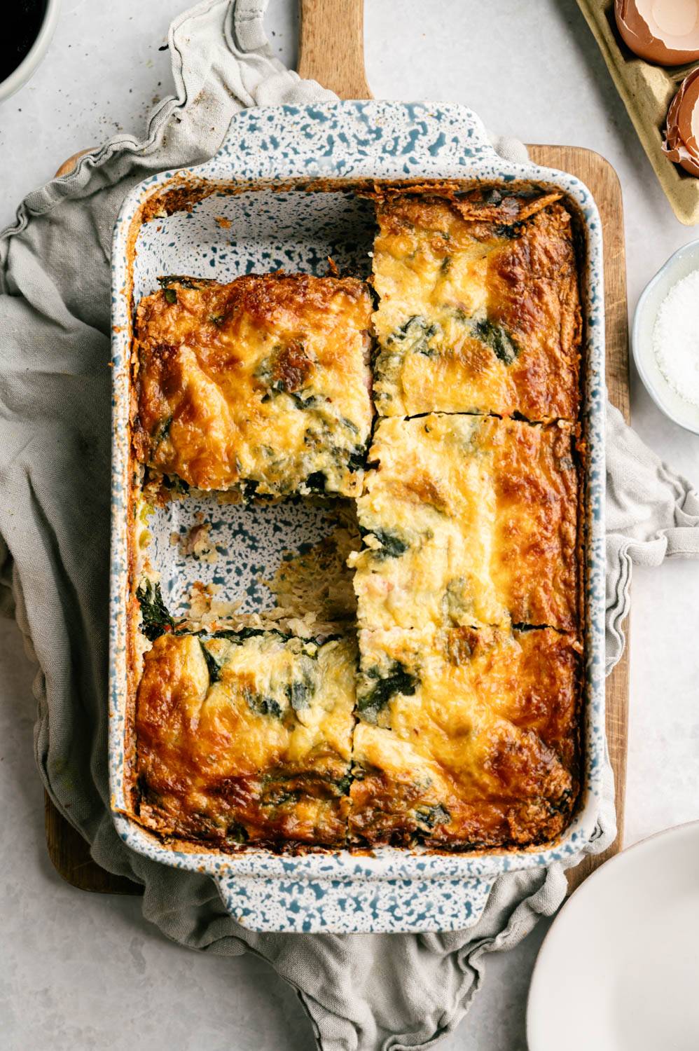 Baked casserole with golden brown cheese cut into cubes in a speckled blue plate on a wooden board covered with a gray cloth napkin.