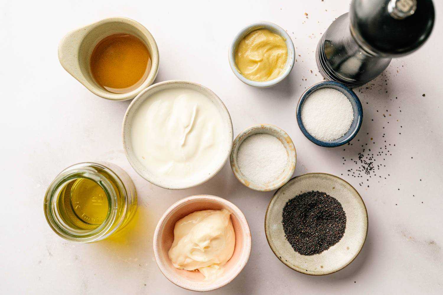 Assorted ingredients for a creamy dressing, including yogurt, mayonnaise, oil, mustard, honey, sugar, salt, poppy seeds, and a pepper grinder on a white surface.