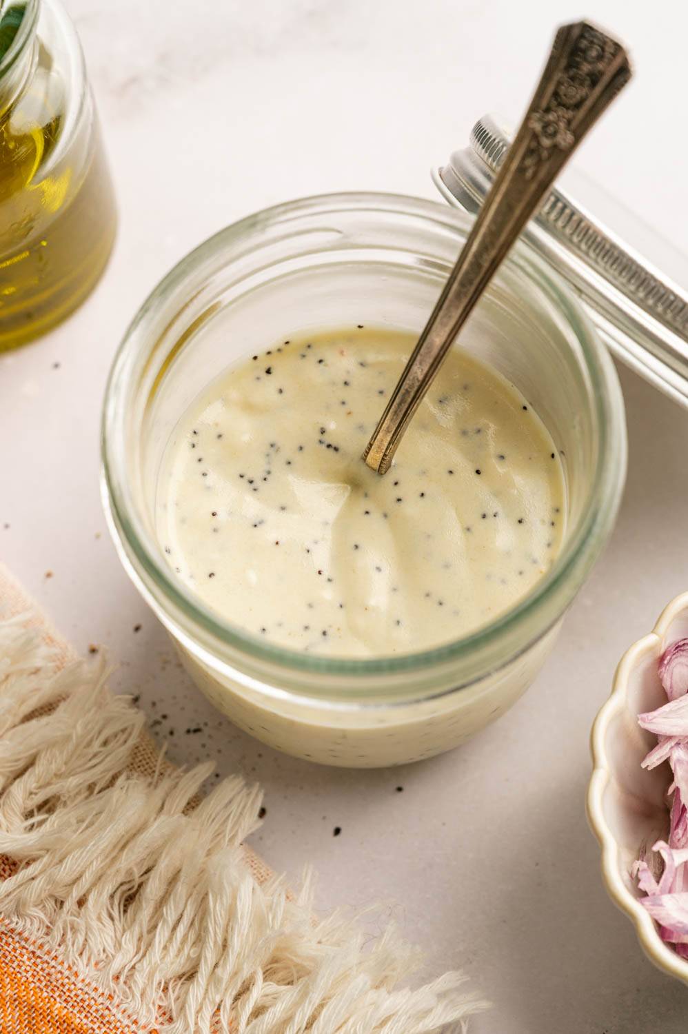 Creamy homemade poppy seed dressing in a glass jar with a spoon, surrounded by olive oil and sliced shallots on a light countertop.