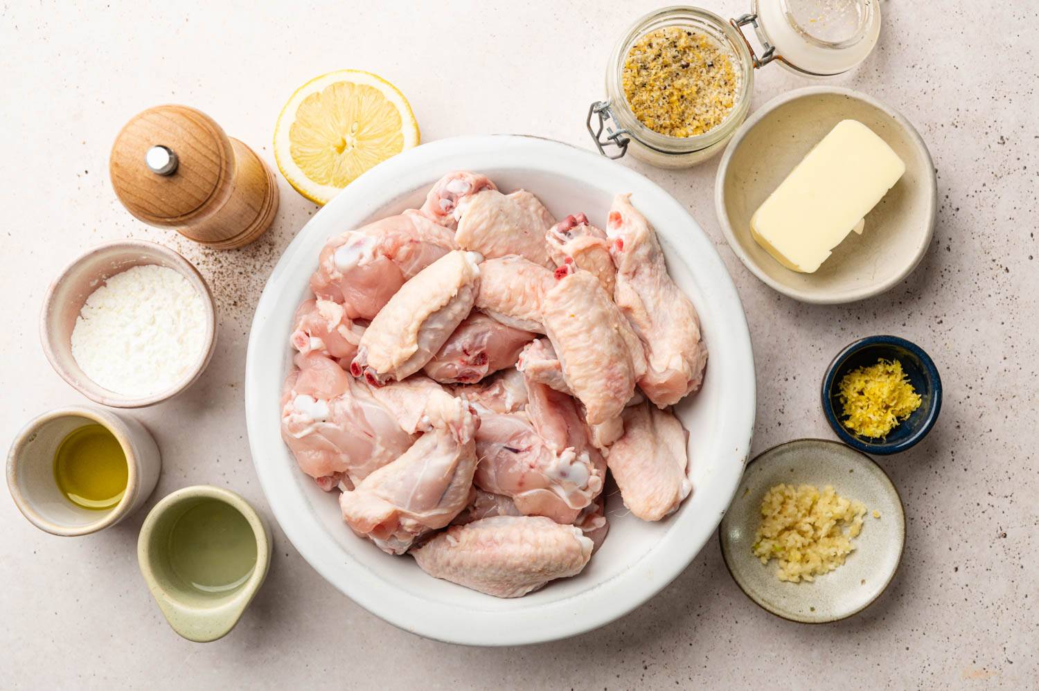 A bowl of raw chicken wings is surrounded by ingredients: butter, lemon, garlic, seasonings, and oil.
