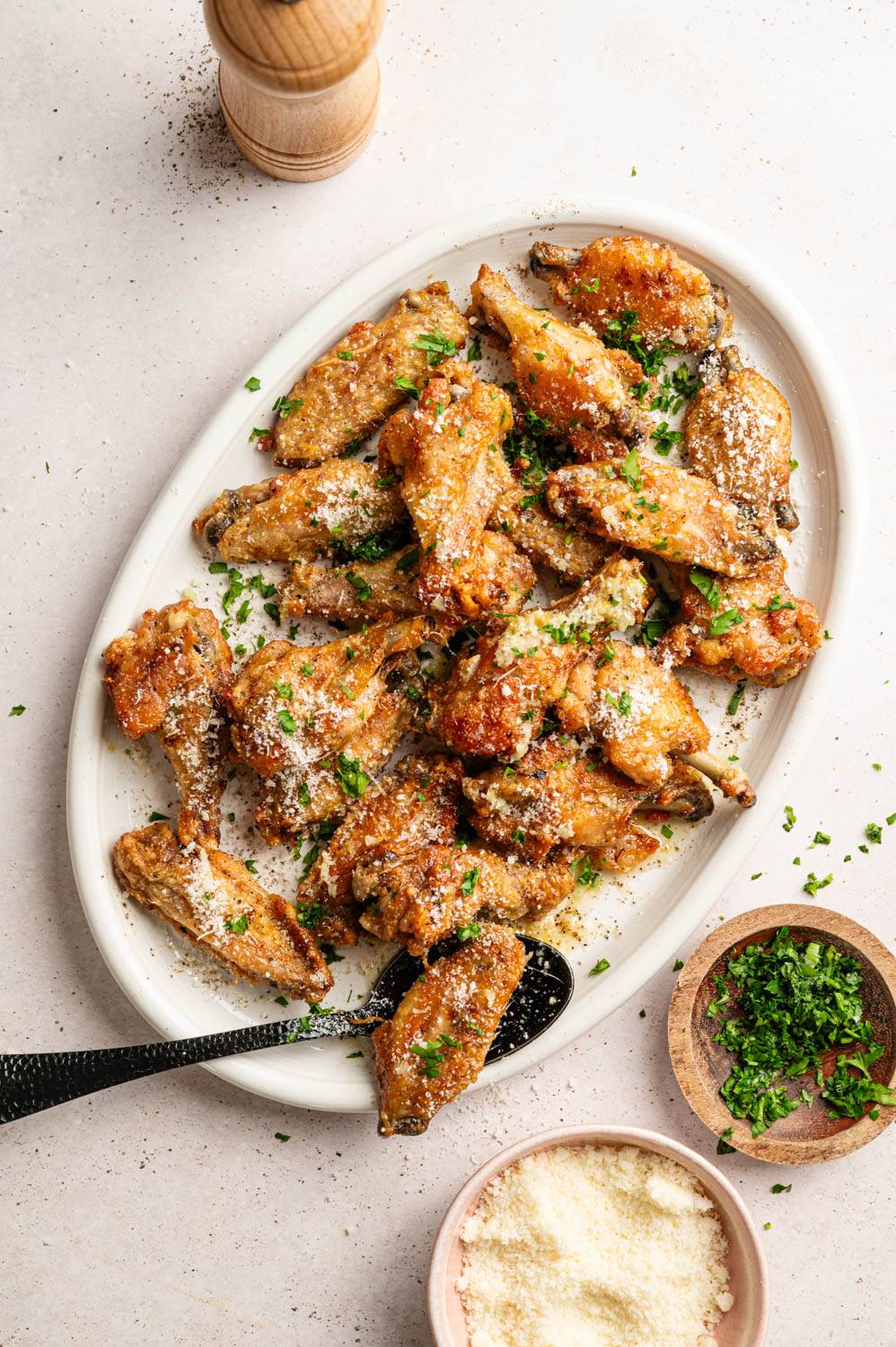 Crispy chicken wings on an oval plate, garnished with parsley and Parmesan. A pepper grinder, parsley, and Parmesan bowls are nearby.