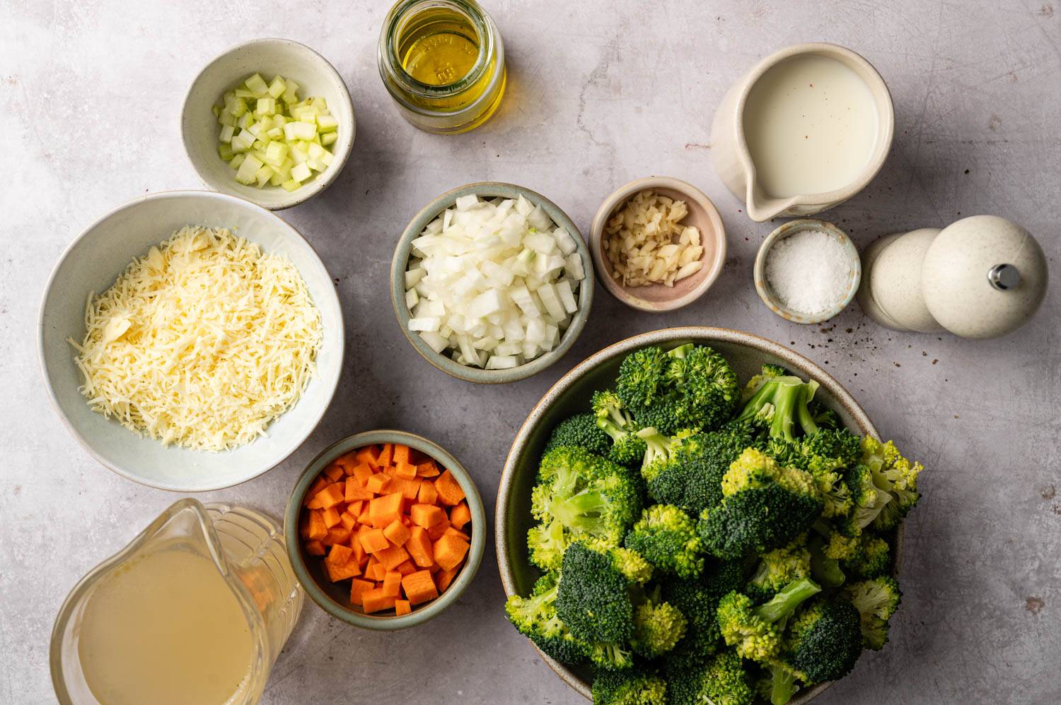Various ingredients for a broccoli soup recipe are laid out on the counter: chopped broccoli, onions, carrots, chopped cheese, garlic, cream, broth, oil and seasonings.